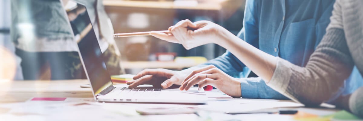 The Cost Benefits of Migrating to the New SQL Server Two workers are looking at a laptop and pointing to it. The photo shows that they are in a busy environment with lots of pens and paper on the table.
