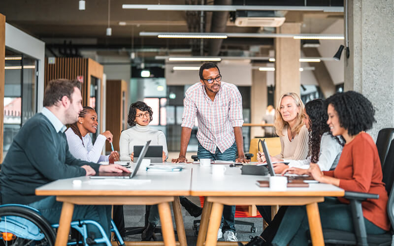 Large team discusses strategy around a conference table in a bright, tech-focused office with glass walls.